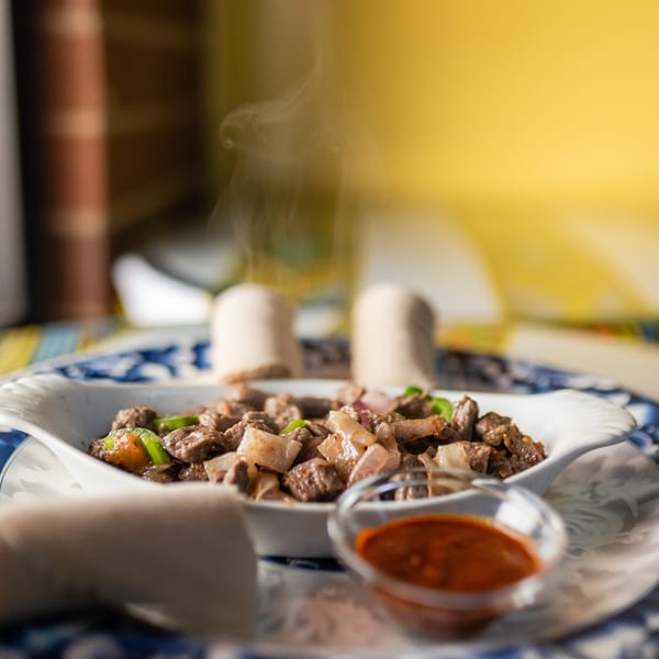 Steaming hot Ethiopian tibs with injera bread and a spicy dipping sauce on a patterned plate.