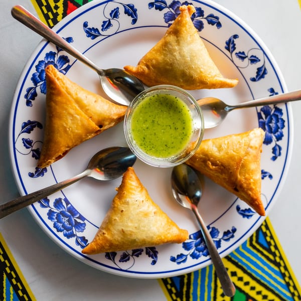 Five golden-brown samosas and a bowl of green chutney on a white plate with blue floral designs.