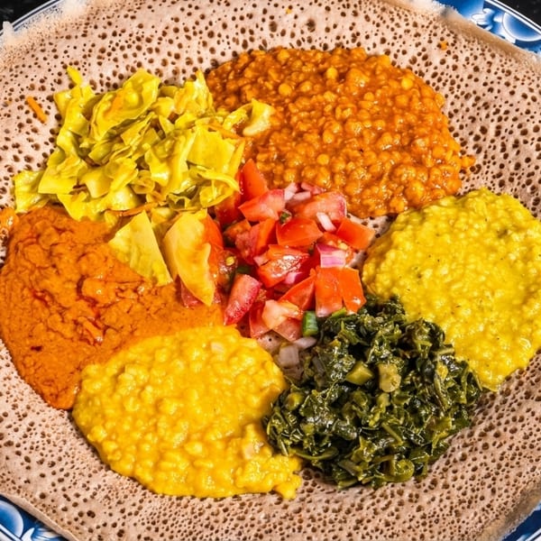 Colorful Ethiopian vegetarian platter with injera, lentils, cabbage, and greens on a blue and white plate.
