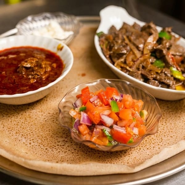 Ethiopian injera bread with three bowls of food: red stew, meat and vegetables, and diced tomatoes.